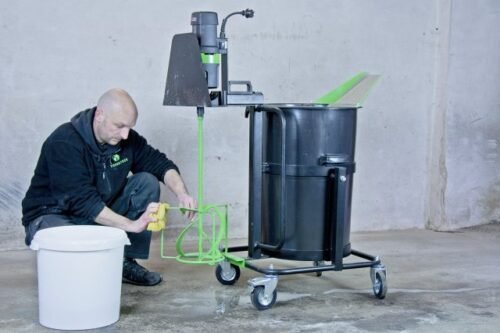 A man in a black hoodie is using a green mixing tool next to a large black container on wheels, with a small white bucket on the floor.